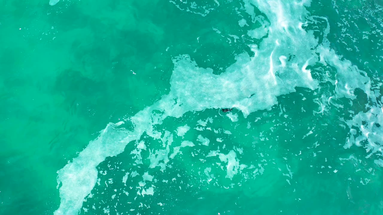 Aerial frame at Marquis Los Cabos, Mexico with the white foam line spread across the center of turquoise water and the tiny surfer shape close to the break, with no recognizable people.