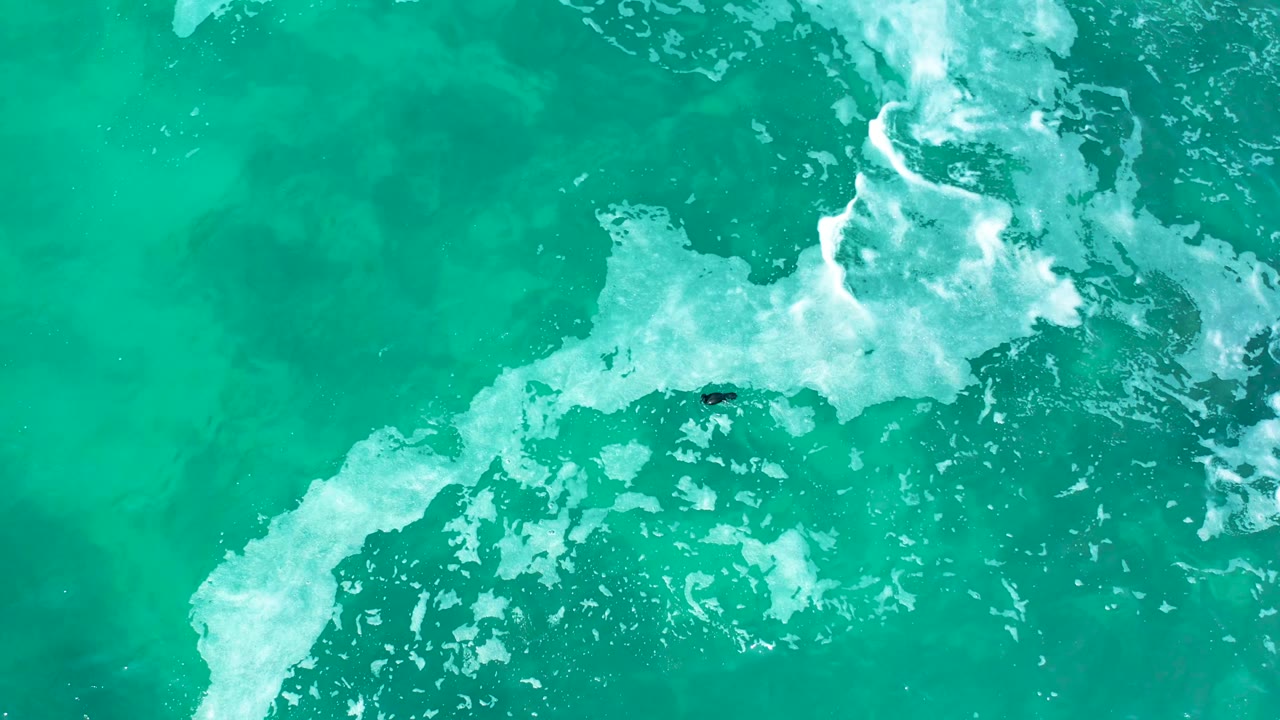 Aerial turquoise sea with diagonal white foam and tiny surfer