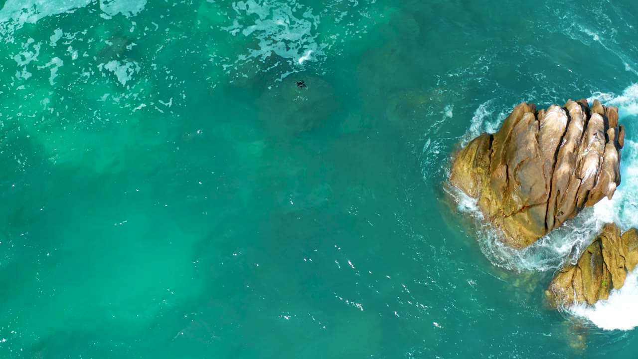 Overhead view at Marquis Los Cabos, Mexico showing turquoise water around a tan rocky outcrop, with a tiny swimmer visible left of the rocks and foam breaking along the edges.