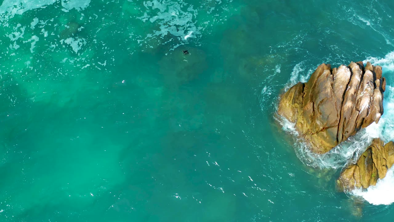 Aerial frame at Marquis Los Cabos, Mexico with the main rock centered right, a smaller rock below it, white foam at both rocks, and a tiny swimmer in open water to the upper left.