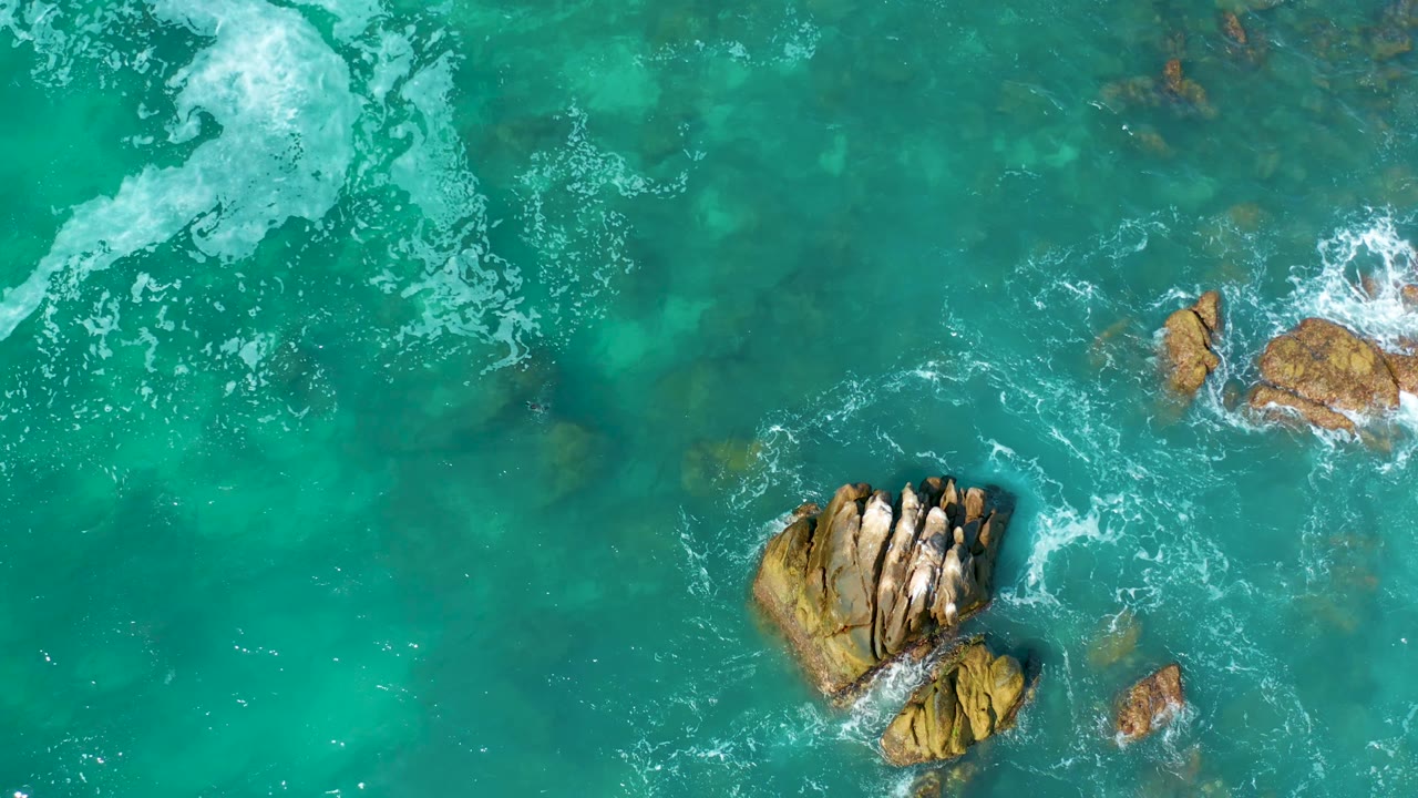 Top-down aerial frame with a large tan rock cluster near the lower left and white surf spreading across turquoise water at Marquis Los Cabos, Mexico, with no visible people.
