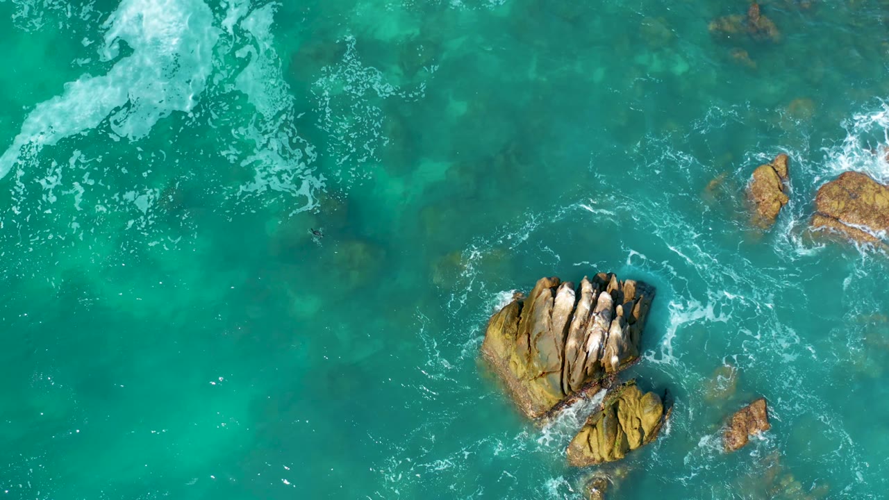 Overhead frame of tan rocky outcrops shifted toward the right side of turquoise water as white foam breaks nearby at Marquis Los Cabos, Mexico, with no visible people.