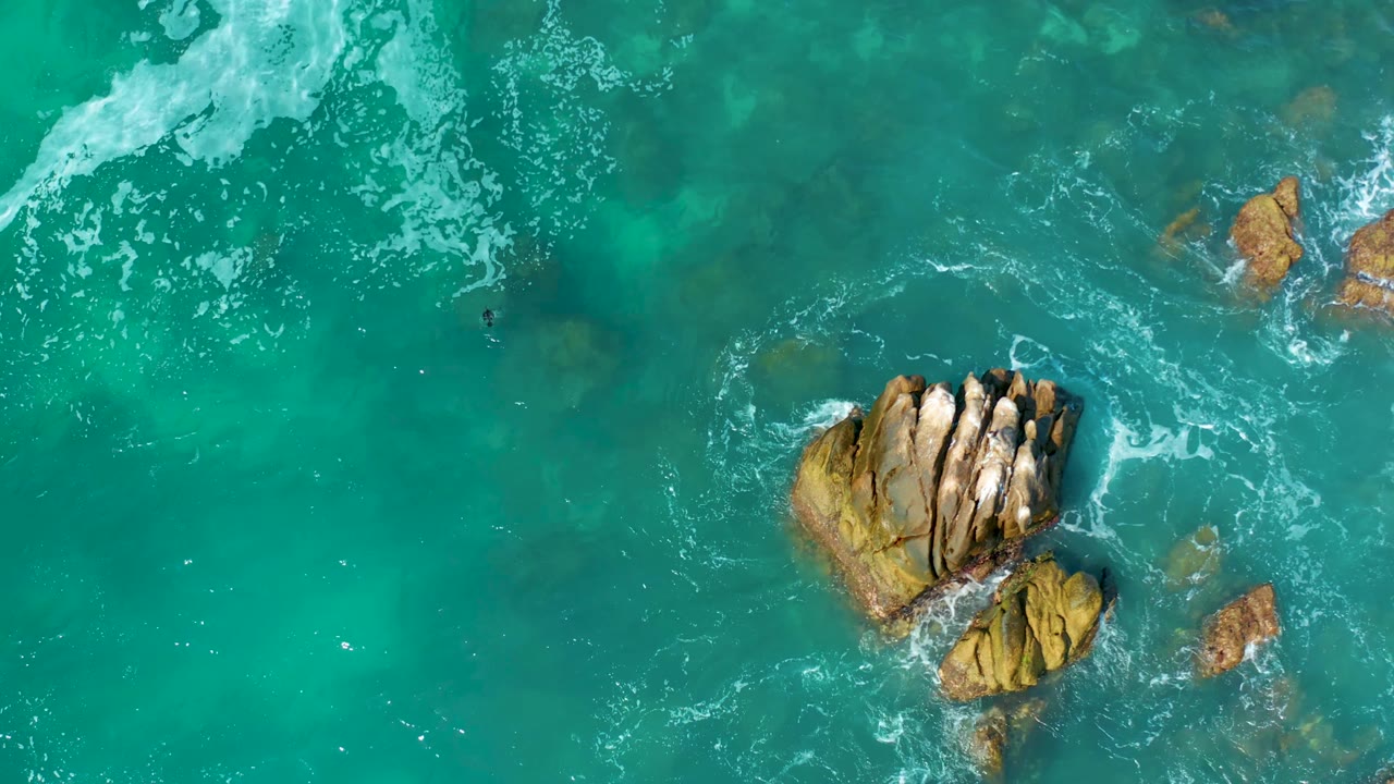 Aerial view of a central tan rock formation with smaller outcrops and white foam curling through bright turquoise water at Marquis Los Cabos, Mexico, with no visible people.