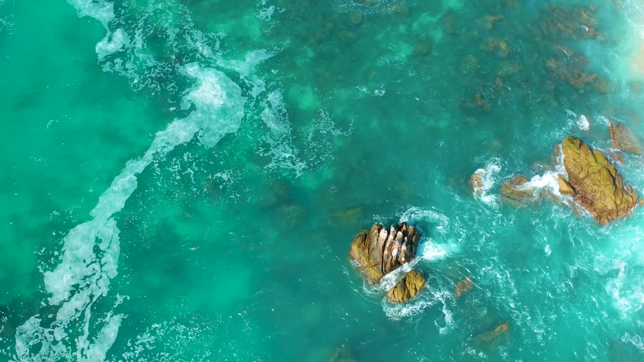Aerial frame from Marquis Los Cabos, Mexico, with a broad white foam trail cutting through green-blue water near exposed tan rocks and no visible people.