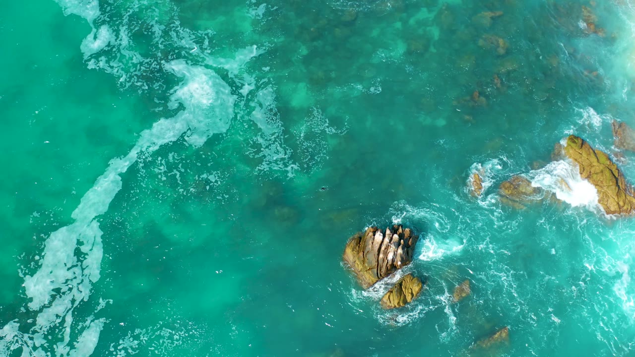 Top-down view at Marquis Los Cabos, Mexico, where white surf breaks over a right-side rock while a central rock cluster sits in clear turquoise water with no visible people.