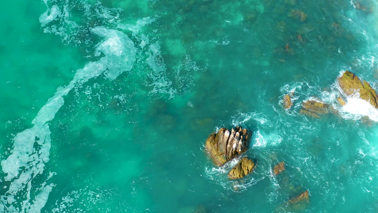Overhead Marquis Los Cabos, Mexico frame with the main rock cluster lower in view, clear teal water around it, and white foam spreading along rocks on the right with no visible people.