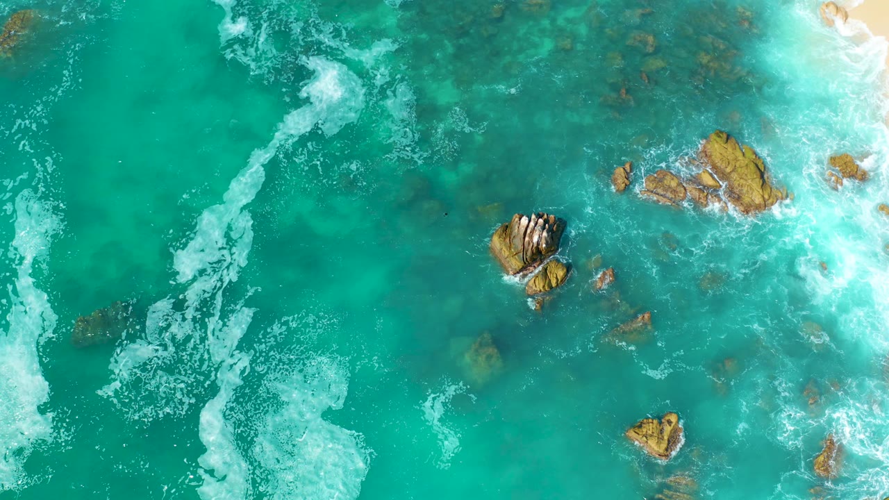 Top-down view at Marquis Los Cabos, Mexico, showing turquoise water, scattered submerged rocks, white foam at left and right, and no visible people.
