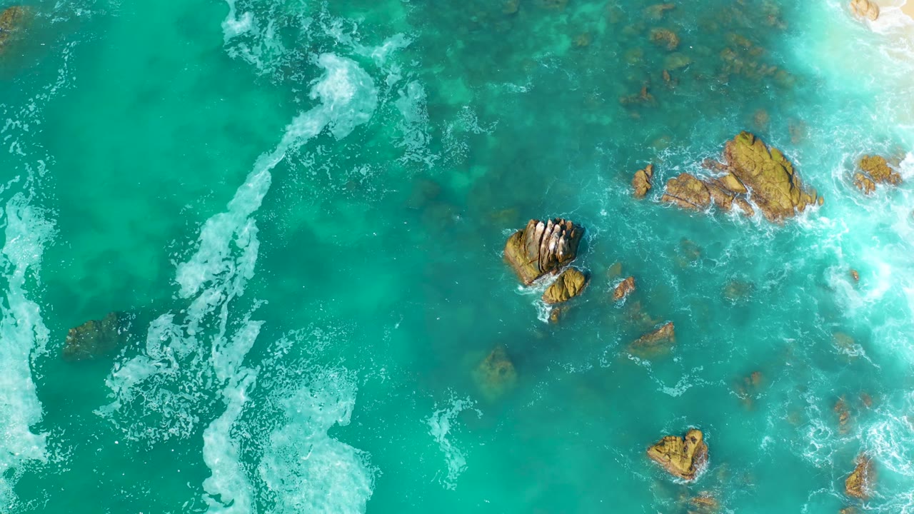 Overhead view at Marquis Los Cabos, Mexico, where white foam breaks across the right side and rocky patches sit under clear turquoise water, with no visible people.