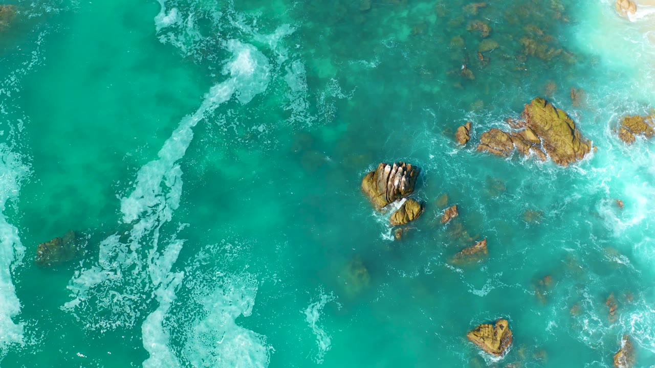 Top-down Marquis Los Cabos, Mexico frame with a stronger diagonal foam streak on the left, brown rocks on the right, and no visible people or boats.