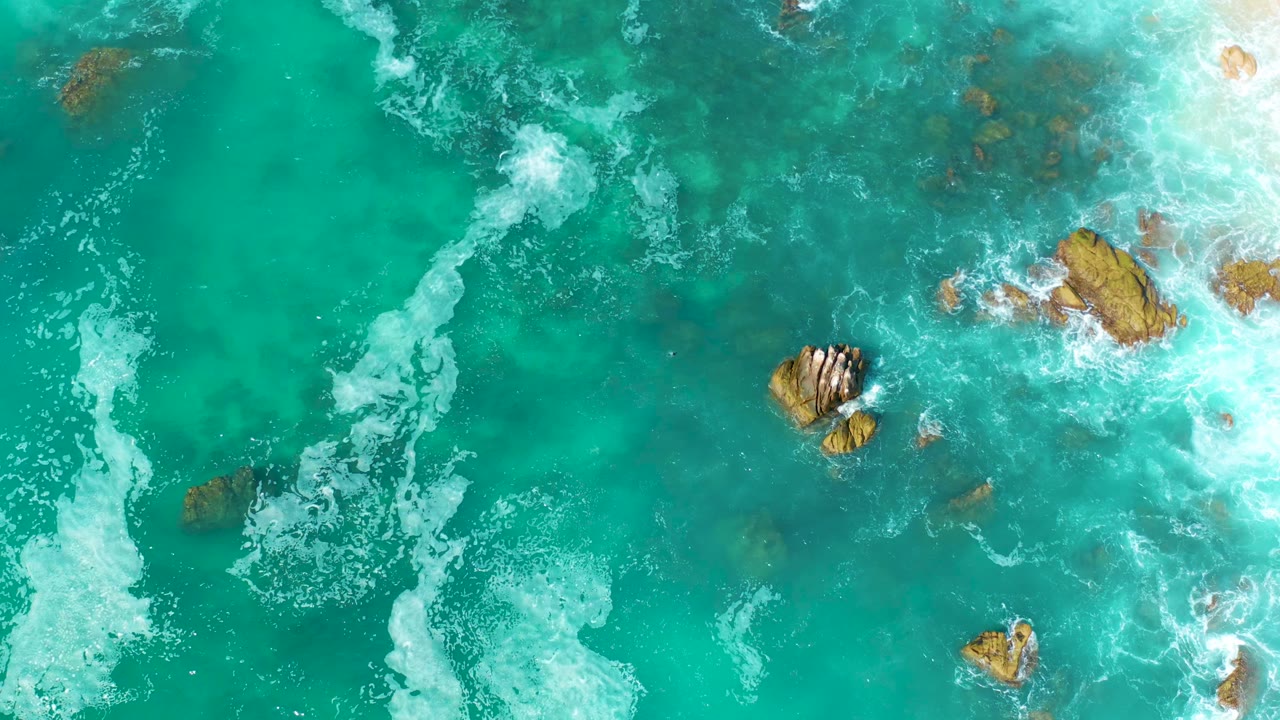 Aerial frame at Marquis Los Cabos, Mexico, with bright turquoise water, a curving foam line on the left, and clustered rocks toward the right center with no visible people.