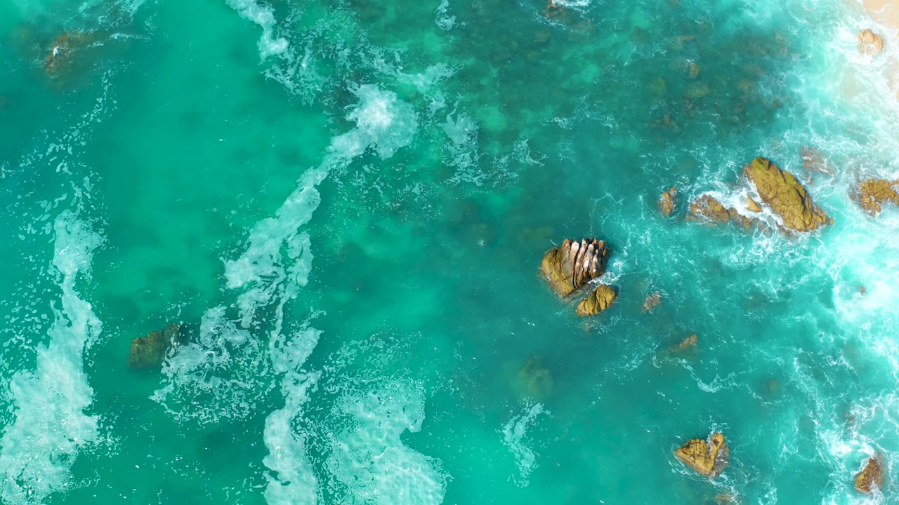 Aerial frame at Marquis Los Cabos, Mexico, showing a central rock cluster, scattered smaller rocks, and bright foam pressing in from the right edge with no visible people.