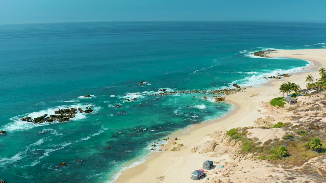 Aerial view of turquoise waves breaking on rocks beside a sandy beach.