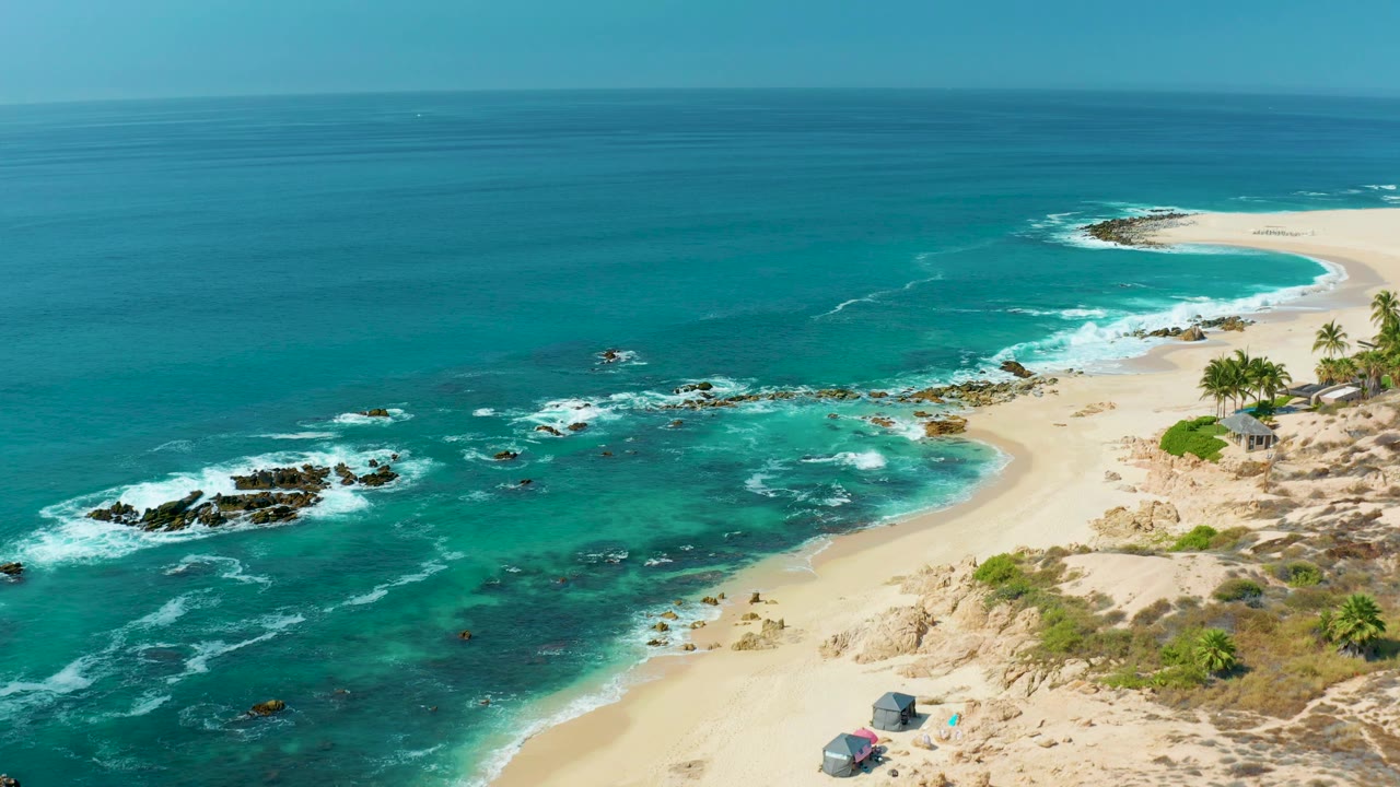 Aerial view at Marquis Los Cabos, Mexico with white foam spreading through shallow turquoise water, dark rocks left of center, sandy beach to the right, and no visible people.