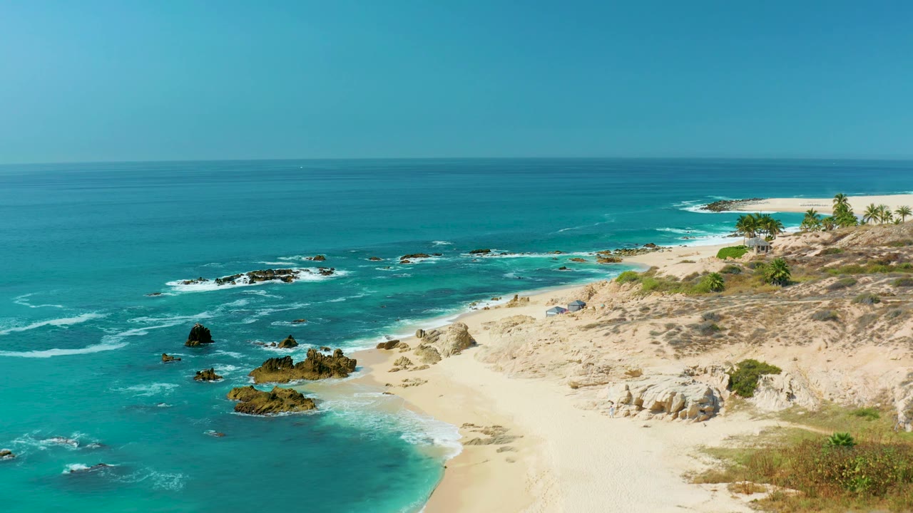 The view shifts along the Marquis Los Cabos shoreline, showing turquoise water, scattered black rocks, pale sand, and no visible people.
