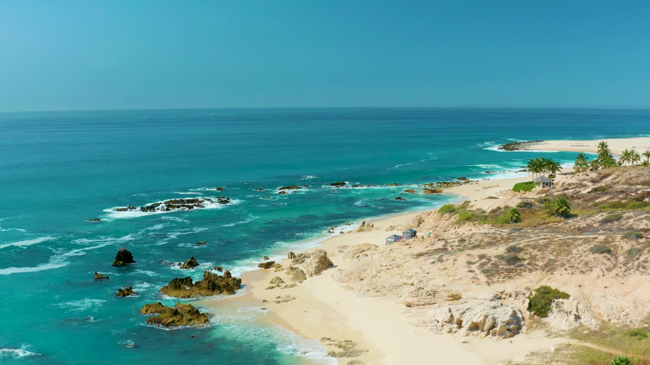A high view over the Marquis Los Cabos coast shows white surf around offshore rocks, a sandy beach, dry scrub on the shore, and no visible people.