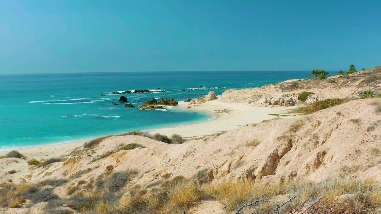 Wide coastal view at Marquis Los Cabos, Mexico, showing pale dunes, scrub, a crescent of empty sand, turquoise ocean, offshore rocks, and no visible people.
