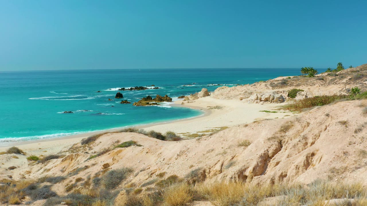 Empty beach cove with turquoise water, dunes, scrub, and rocks offshore.
