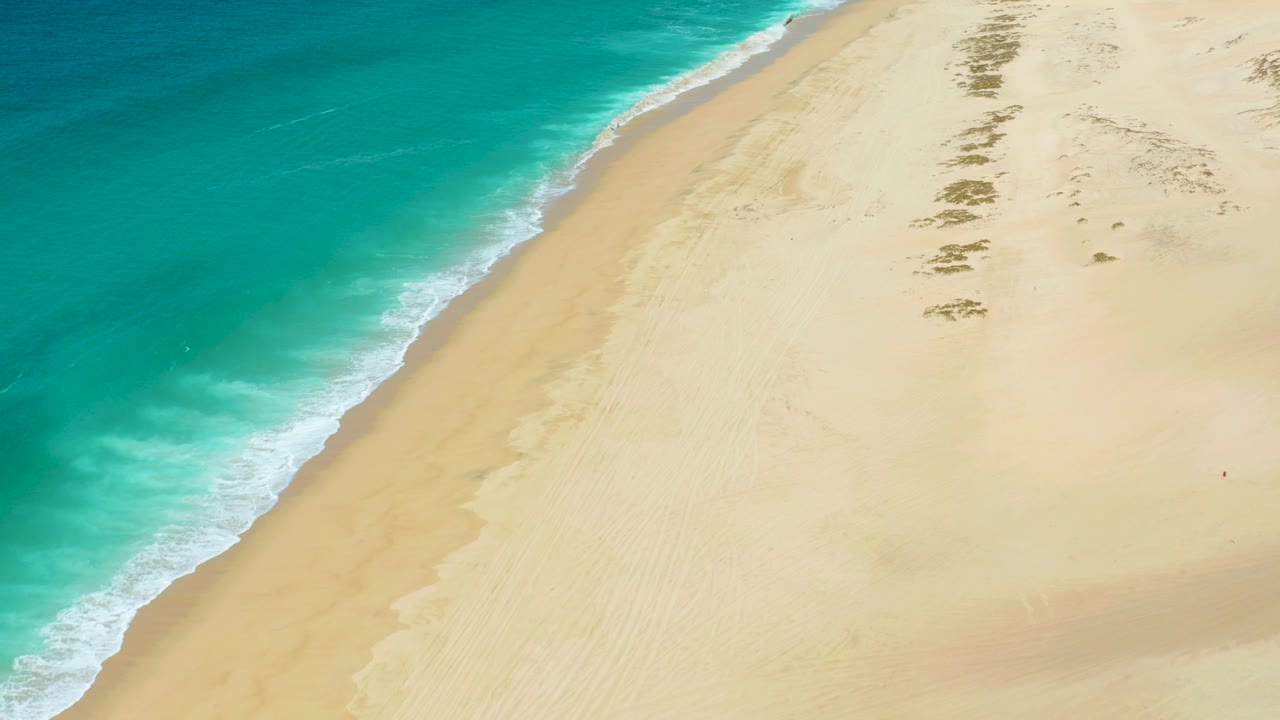 Aerial view of turquoise water rolling onto pale sand at La Laguna Beach Los Cabos, with scattered dark beach patches and no visible people.
