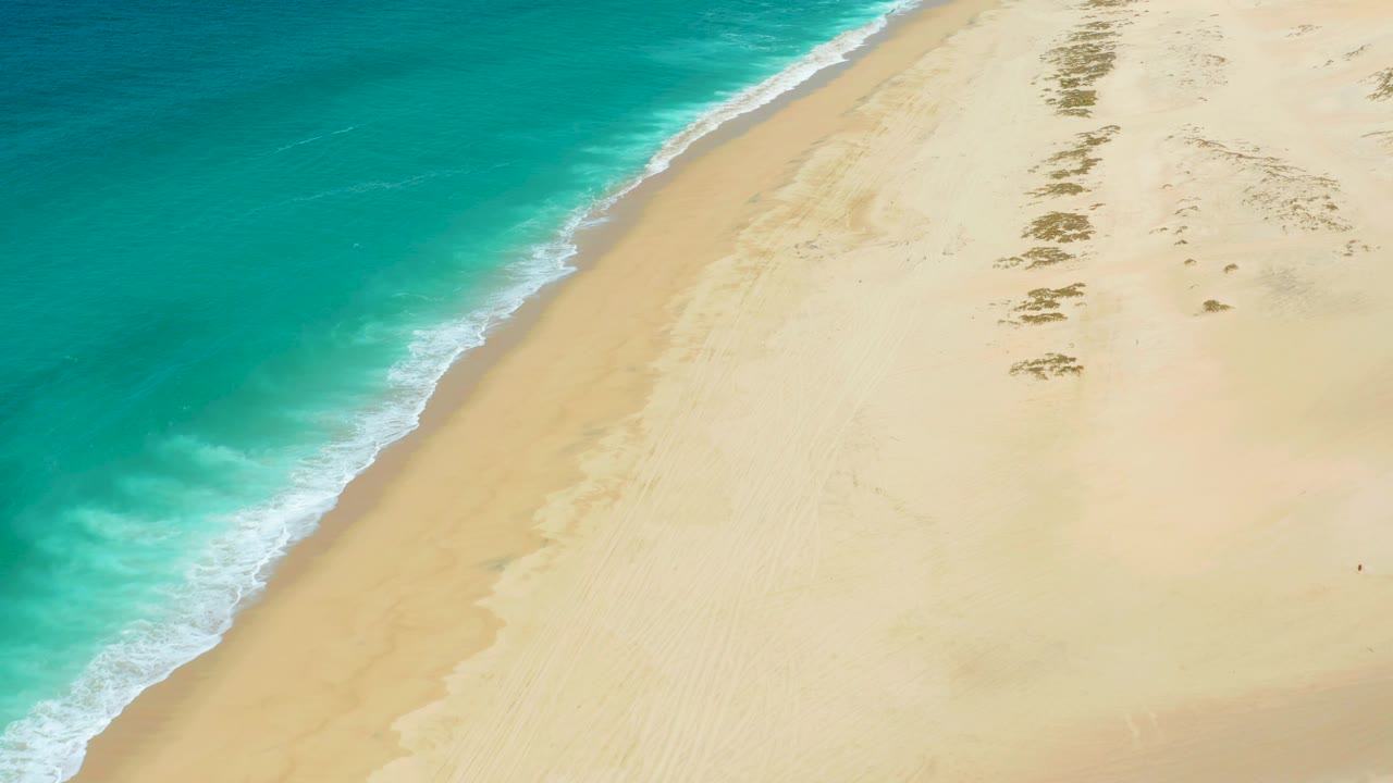 Aerial view of a small wave wash spreading onto the sand at La Laguna Beach Los Cabos, with turquoise water on the left and no visible people.