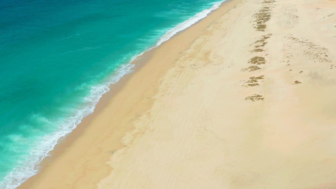 Aerial beach frame from La Laguna Beach Los Cabos showing empty pale sand, turquoise water, a foamy diagonal shoreline, and scattered dark patches farther up the beach.