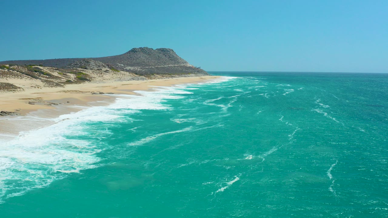 Aerial view of La Laguna Beach Los Cabos with turquoise water, white surf on pale sand, scrubby dunes, a rocky hill, blue sky, and no visible people.