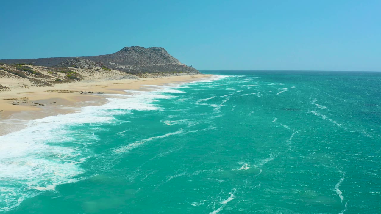 La Laguna Beach Los Cabos seen from above with surf washing toward the beach, a broad turquoise foreground, arid dunes behind the sand, and no visible people.
