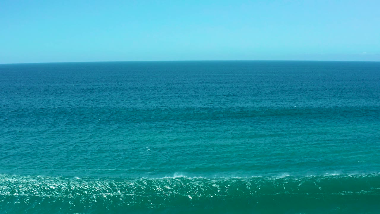 Aerial frame over La Laguna Beach Los Cabos water with turquoise foreground, darker blue midground swell, and a clear horizon, no visible people.
