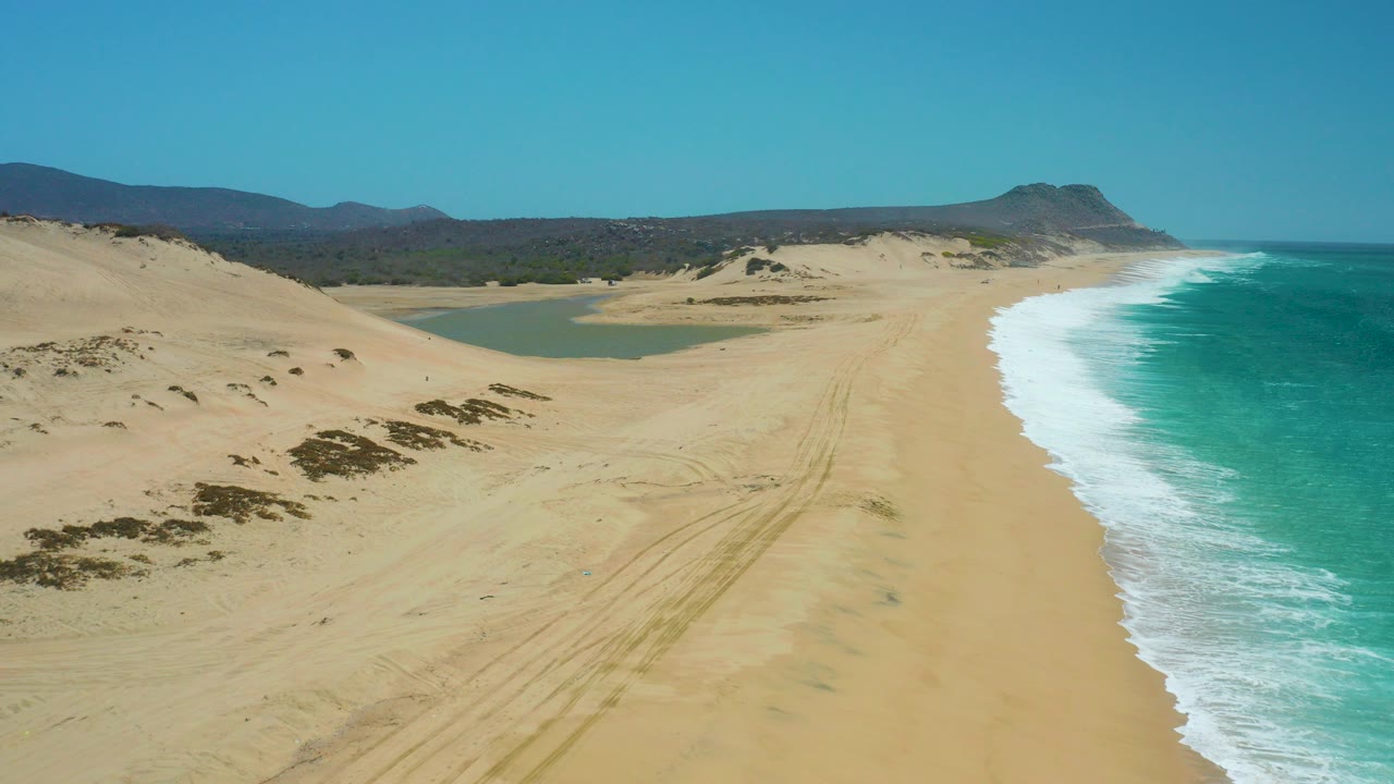 Aerial view over La Laguna Beach with the lagoon centered left, tire-like sand tracks on the beach, white surf along the right edge, and no visible people.