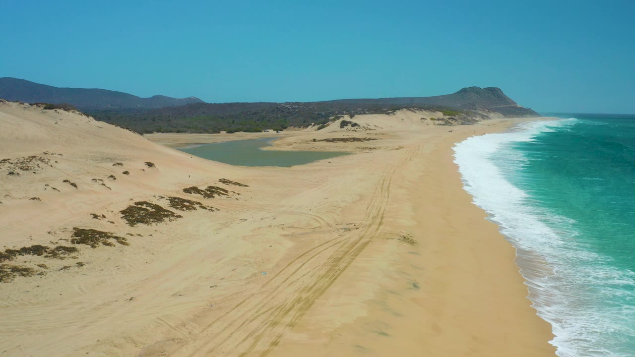 Elevated La Laguna Beach frame with dunes sweeping in from the left, a green-blue lagoon behind the beach, ocean foam on the right, distant hills, and no visible people.