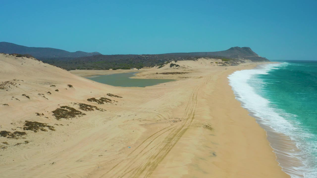 Sandy dunes beside lagoon and ocean waves under clear sky