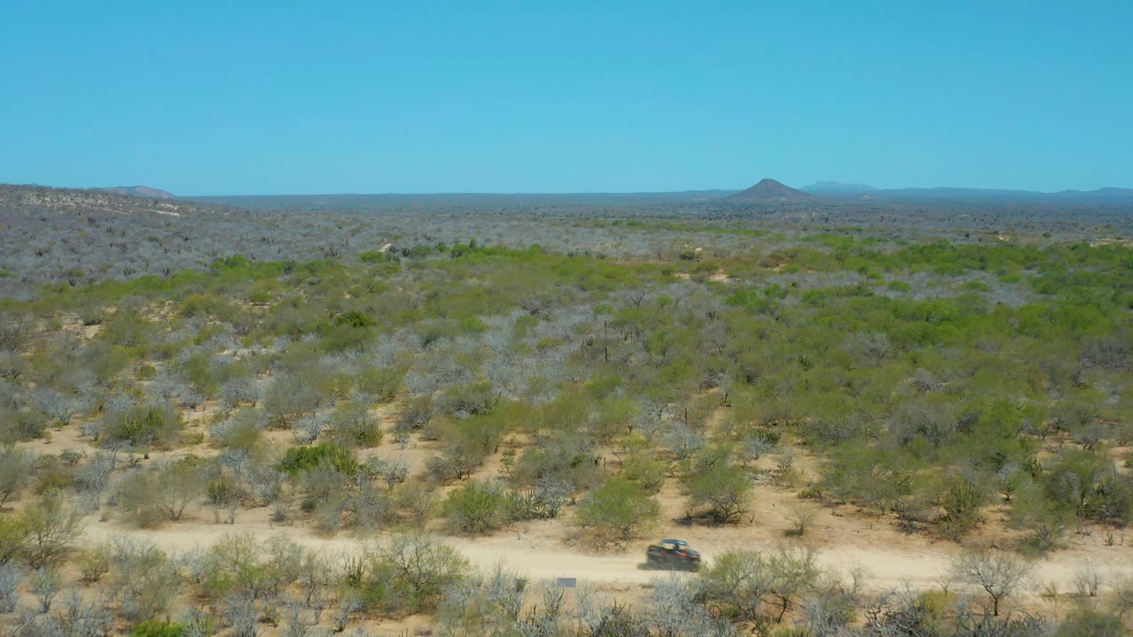 The vehicle sits lower in the elevated La Laguna Beach Los Cabos frame on a pale dirt road, with gray-green desert brush, cacti, distant hills, and no visible people.