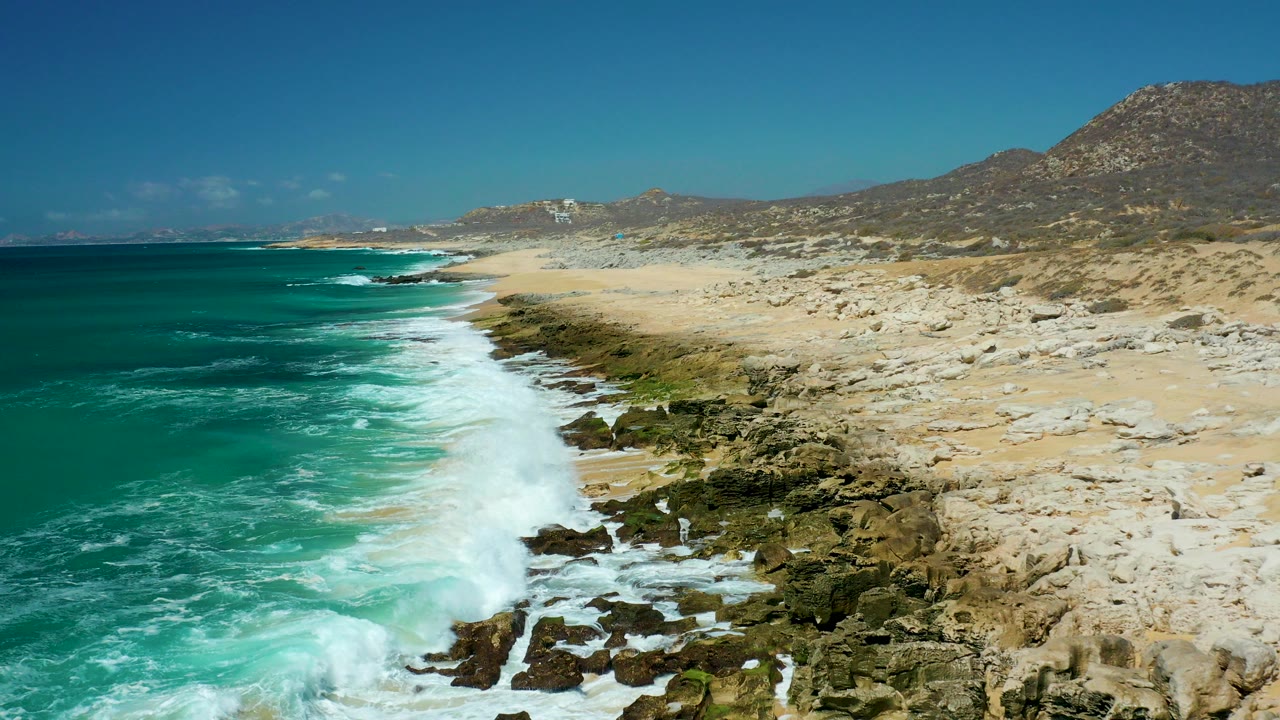Aerial view of La Laguna Beach Los Cabos with turquoise water, white surf, pale rocky shore, distant dry hills, and no visible people.