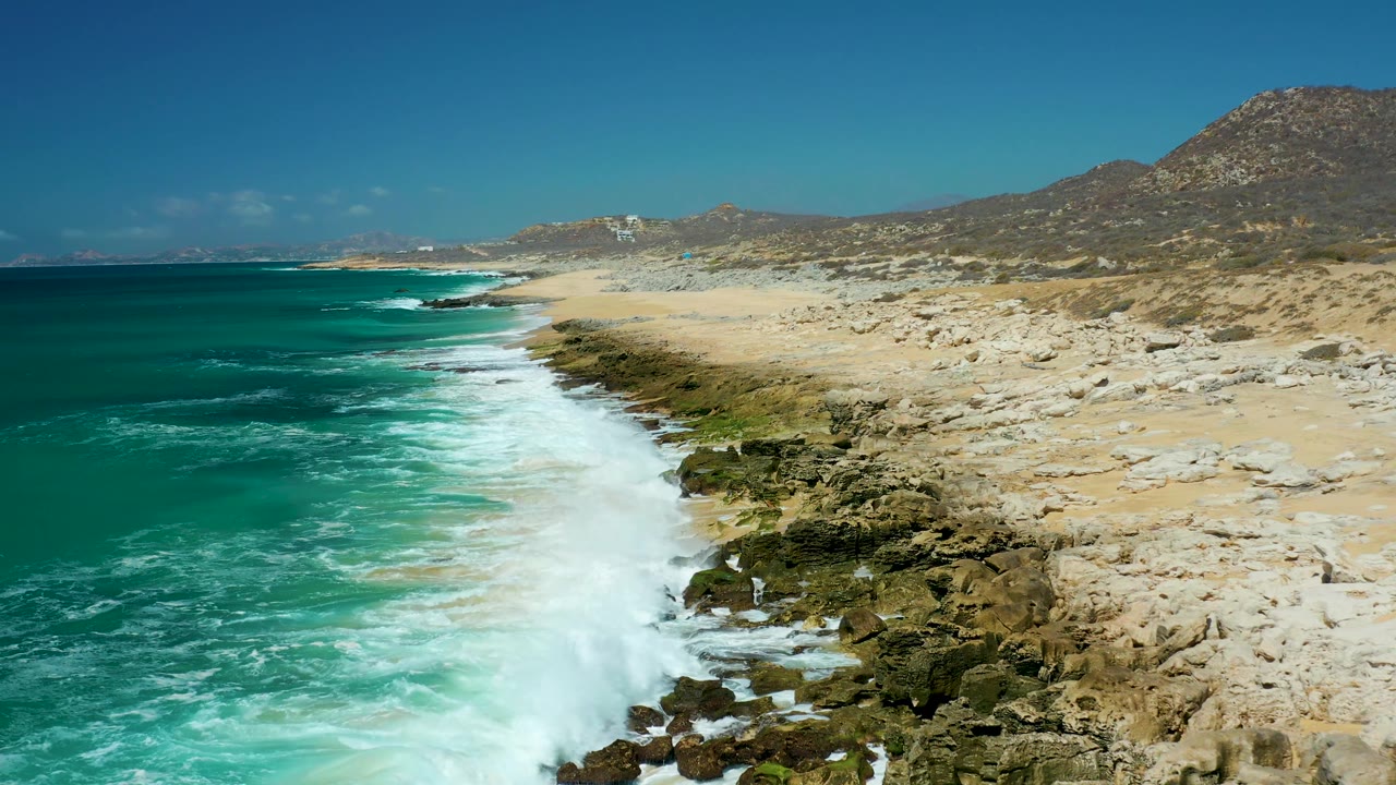 Aerial frame of La Laguna Beach Los Cabos where a larger white wave breaks against dark rocks beside turquoise water, pale sand, dry hills, and no visible people.