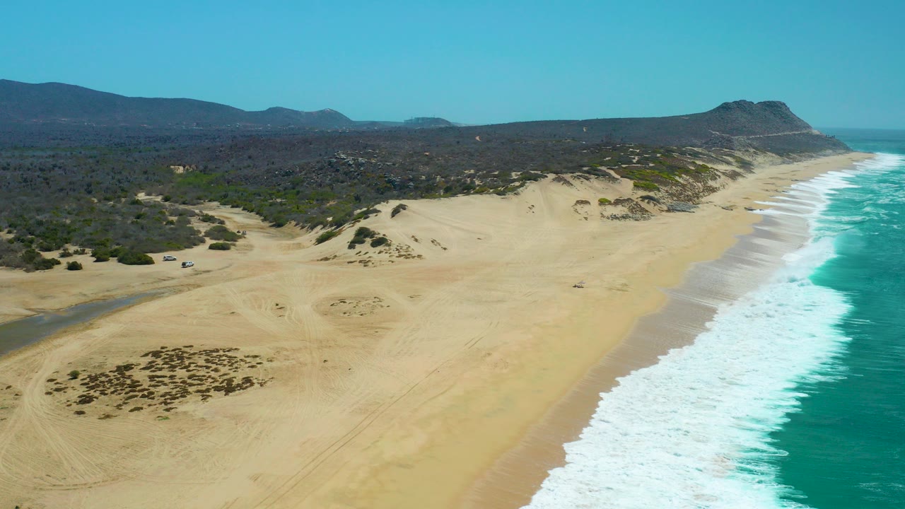 Wide aerial frame at La Laguna Beach Los Cabos showing the surf line slightly farther along the beach, with dune ridges, scrubland, distant hills, clear sky, and no visible people.