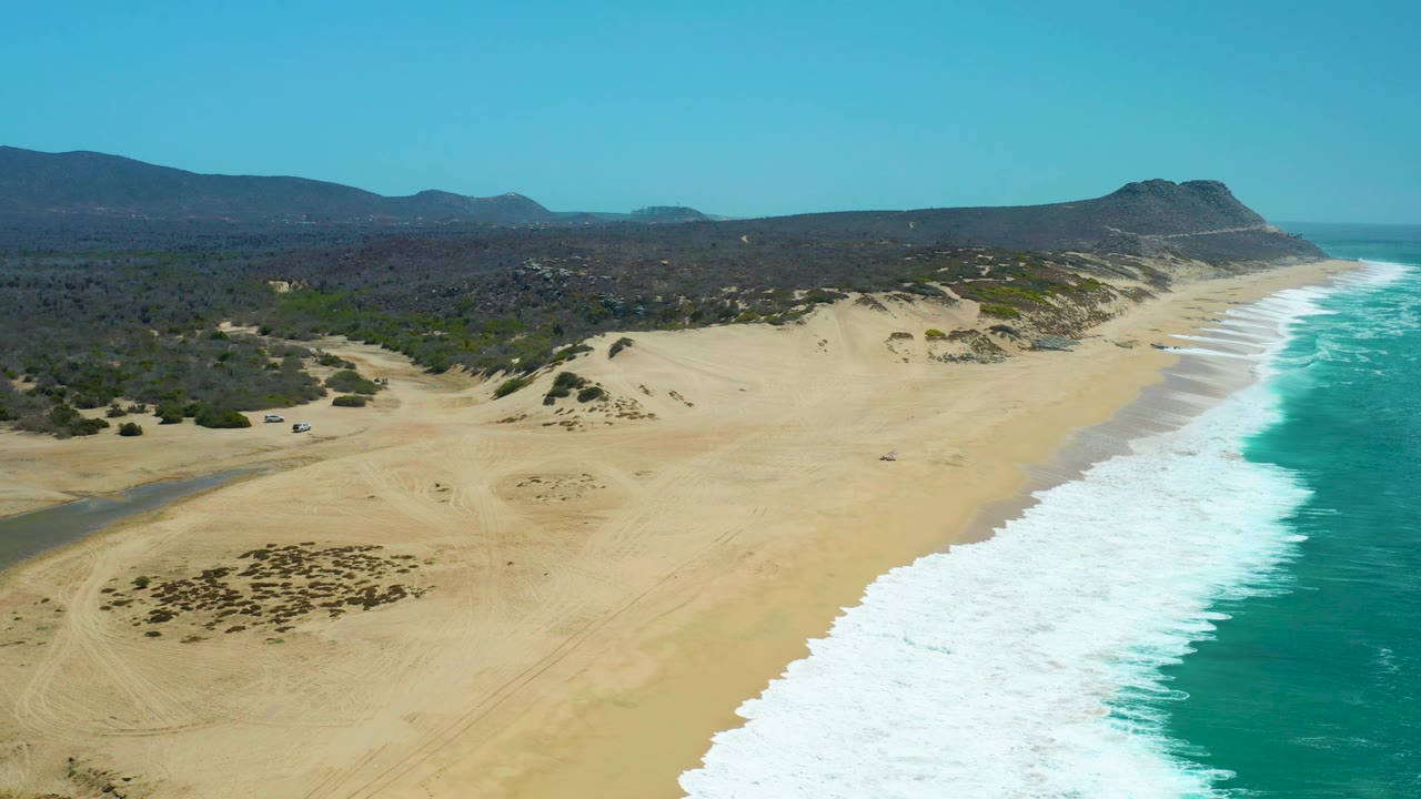 Aerial frame over La Laguna Beach Los Cabos with darker dune vegetation patches in the lower left, bright surf running along the right side, inland hills across the horizon, and no visible people.