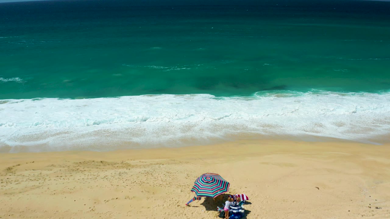 Aerial view of La Laguna Beach Los Cabos with turquoise water, white surf, pale sand, a striped umbrella, and beachgoers visible near the lower edge.