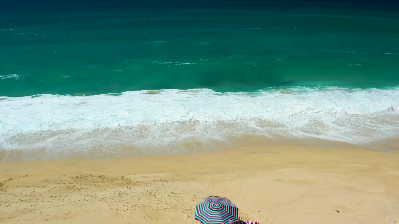 Aerial view of La Laguna Beach Los Cabos with the striped umbrella near the lower right, beachgoers partly visible, and foamy surf stretching across the shore.