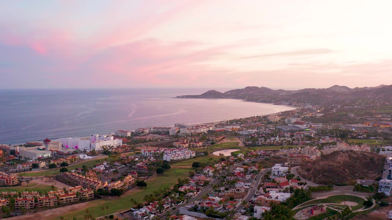 Opening aerial frame over San Jose Del Cabo, Mexico, showing the ocean, curved bay, low-rise town, green areas, hills, and pink dusk clouds with no visible people.