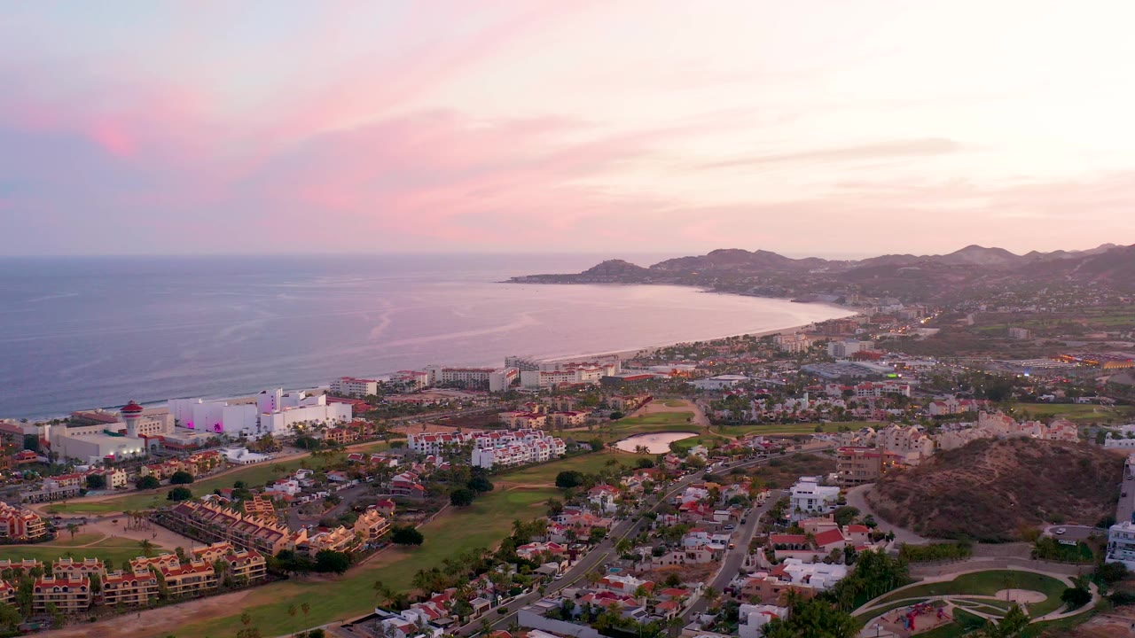 A wide San Jose Del Cabo frame shows the ocean horizon, smooth bay curve, dense town blocks, a hillside at right, and pink cloud bands with no visible people.