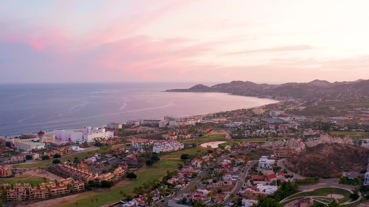 Aerial view of ocean, coastal buildings, green spaces, hillside, and soft pink sky.