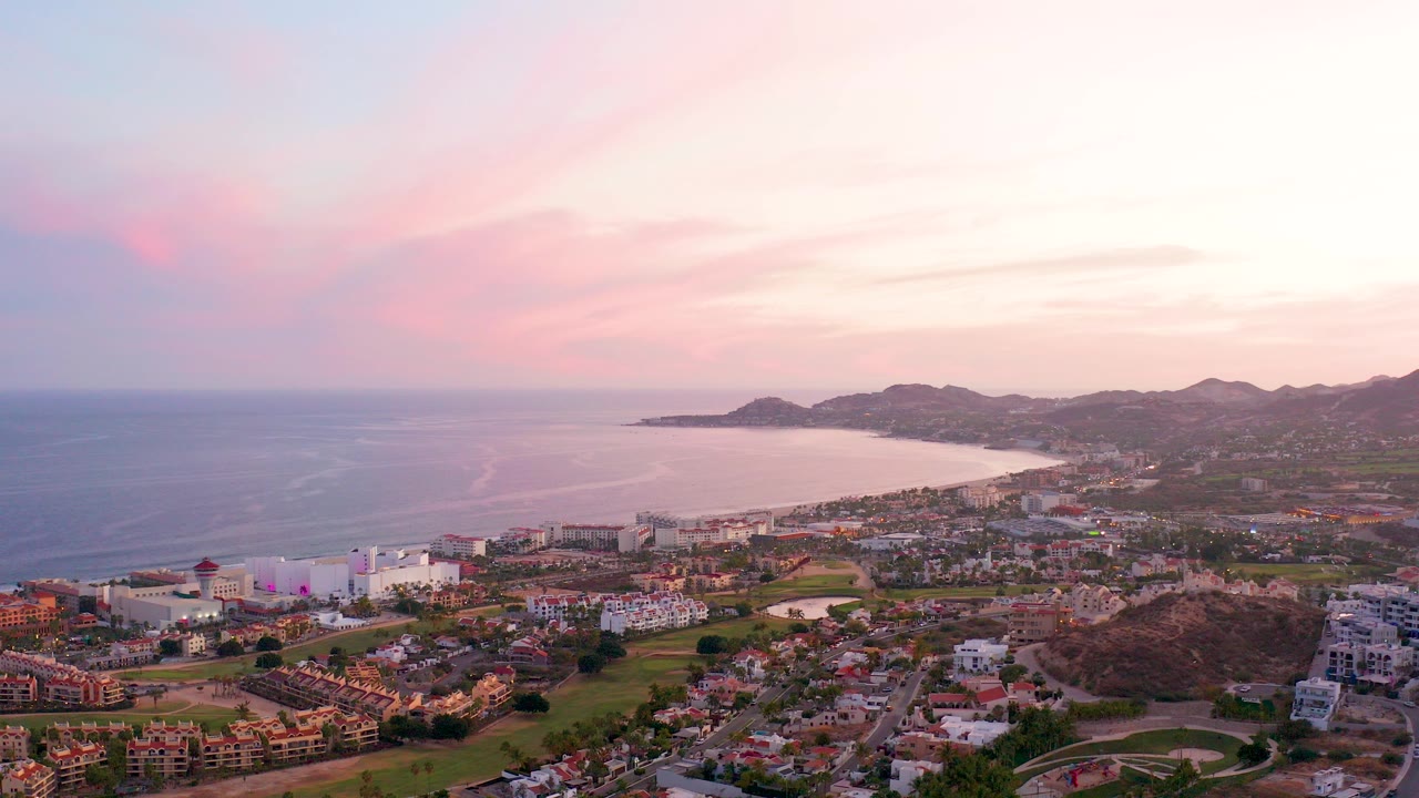 San Jose del Cabo aerial view at 0:00 with ocean on the left, a curved beach and city below, distant mountains, pink sunset clouds, and no visible people.