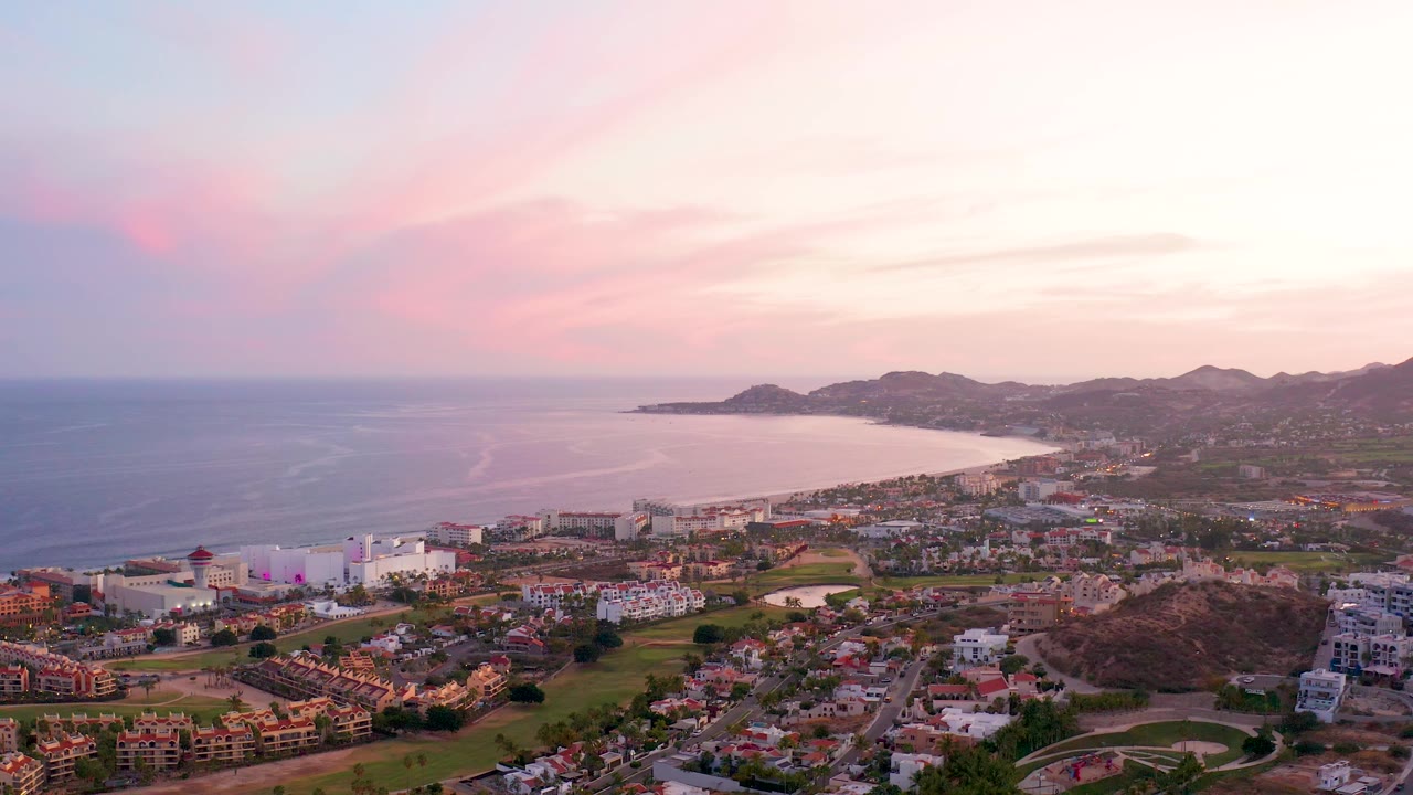 San Jose del Cabo aerial frame at 0:02 with more open ocean visible left of the bay, beachside buildings near the shore, hillside homes below, and no visible people.