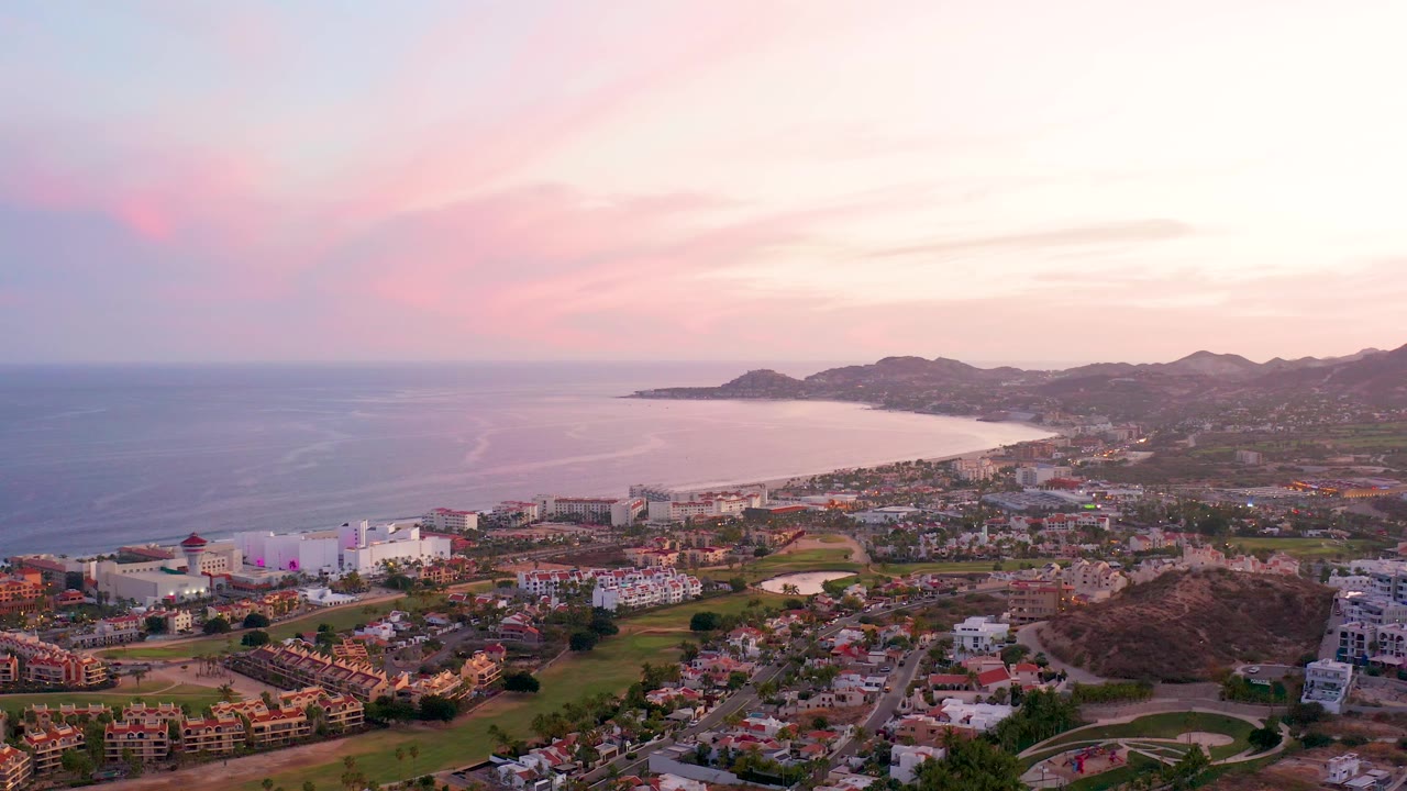 Elevated view of coastline, ocean, city blocks, hills, and pastel sunset sky.