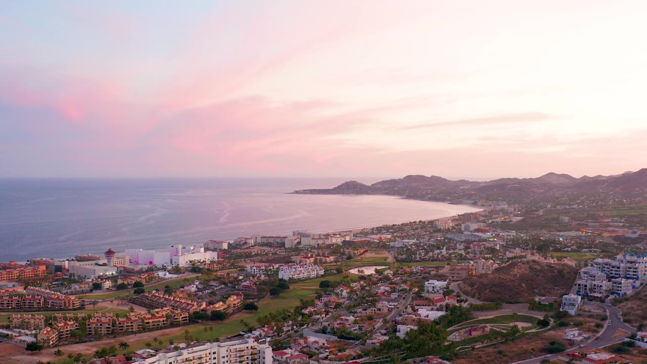 San Jose del Cabo aerial view at sunset with ocean on the left, a curved bay, hillside buildings below, pink clouds, and no visible people.