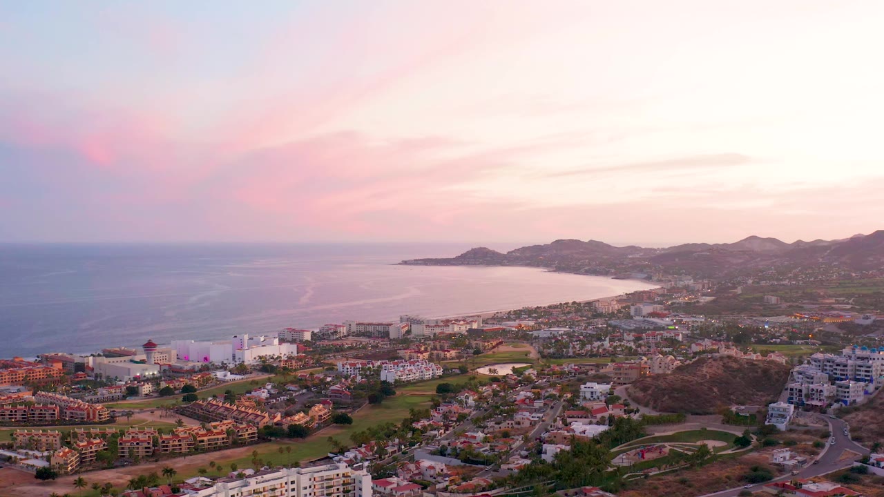 Coastal town and bay under pink sunset clouds with hills beyond.
