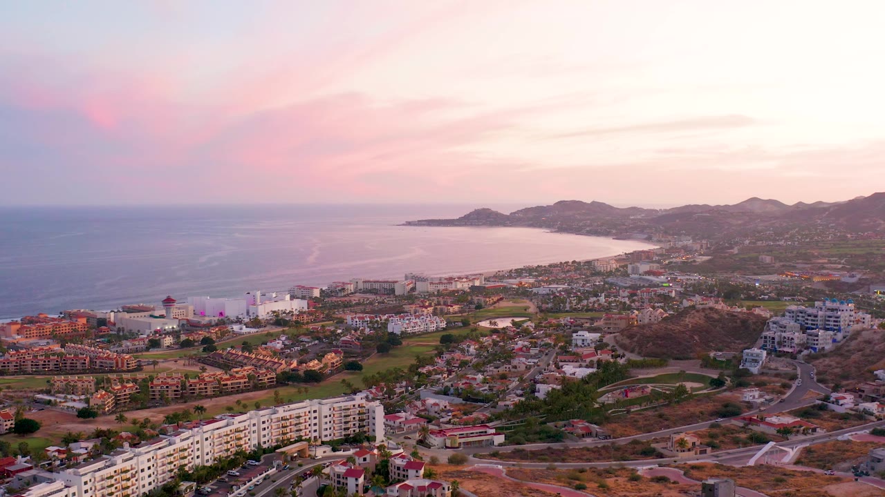 Wide aerial over San Jose del Cabo with apartments and houses in the lower frame, the bay and open ocean stretching left, distant hills on the horizon, and no visible people.