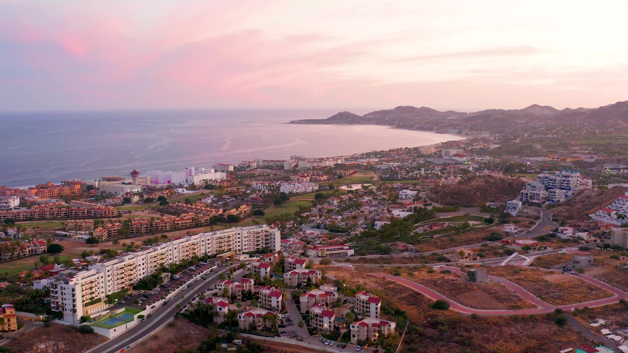 Aerial view over San Jose del Cabo with residential buildings, a curving road, the ocean and bay beyond town, pink dusk clouds, and no visible people.