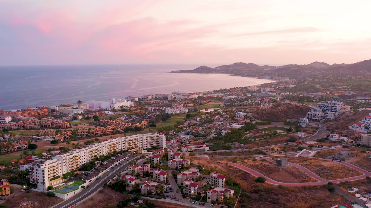 San Jose del Cabo appears from above with apartment blocks and homes in the foreground, small vehicles on the road, the bay in the distance, and no visible people.