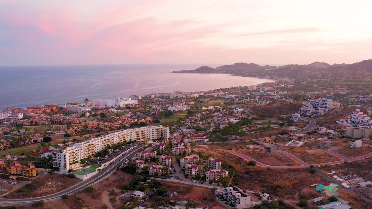 Opening aerial frame over San Jose Del Cabo with residential buildings, curved roads, ocean, distant hills, pink sunset clouds, and no visible people.