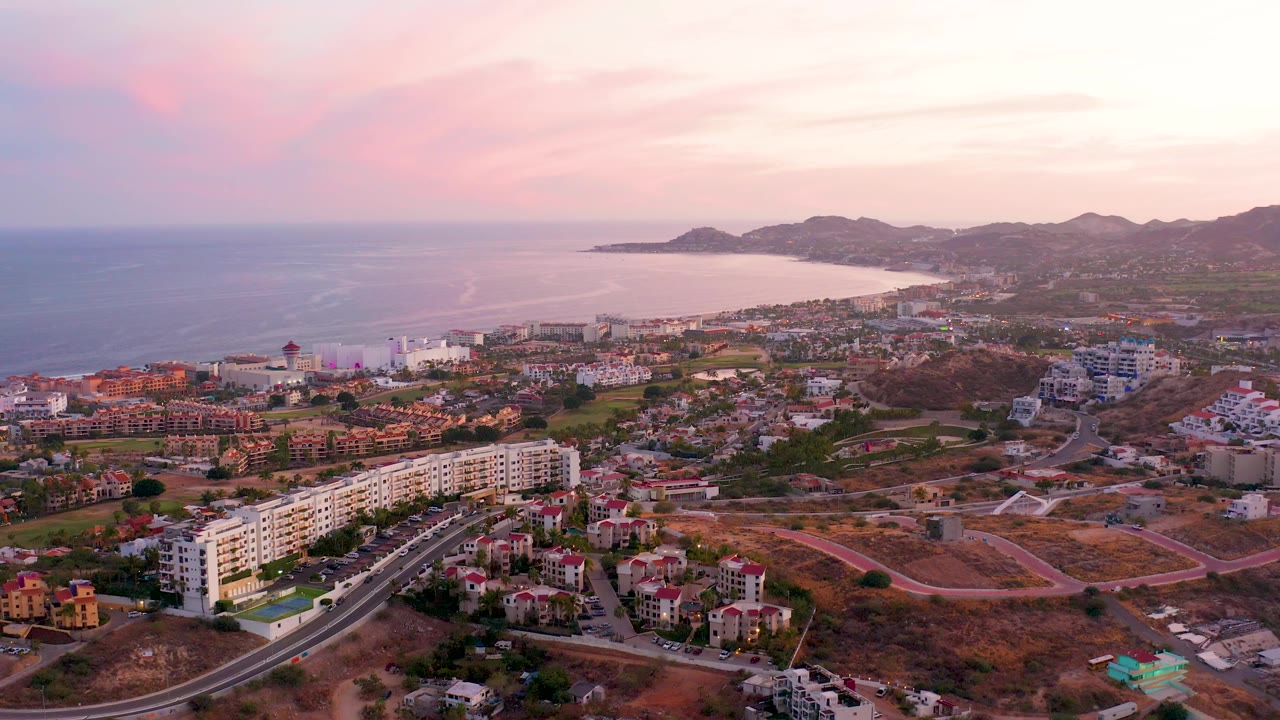 Sunset aerial over coastal buildings, dry lots, roads, ocean, and hills.